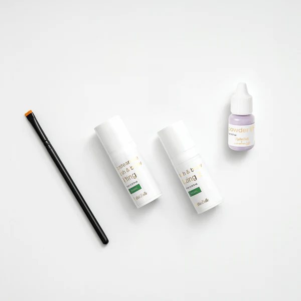 Assorted cosmetic bottles and a small lavender dropper bottle on a white surface with a black makeup brush on the left.